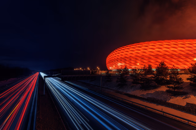 A football stadium glowing red at night beside light trails on a road